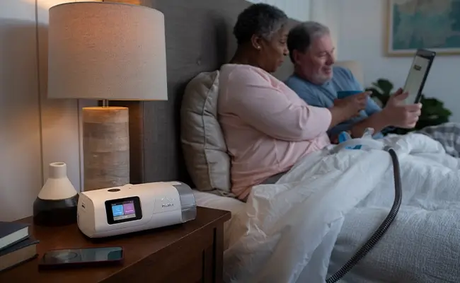 Un couple au lit regardant une tablette électronique. La femme a un masque pour AirCurve 11 ASV PaceWave sur ses genoux. Le masque est relié à l'appareil qui se trouve sur sa table de chevet.